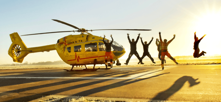 Crew jumping next to yellow, EAAA helicopter at sunset creating shadows on the ground